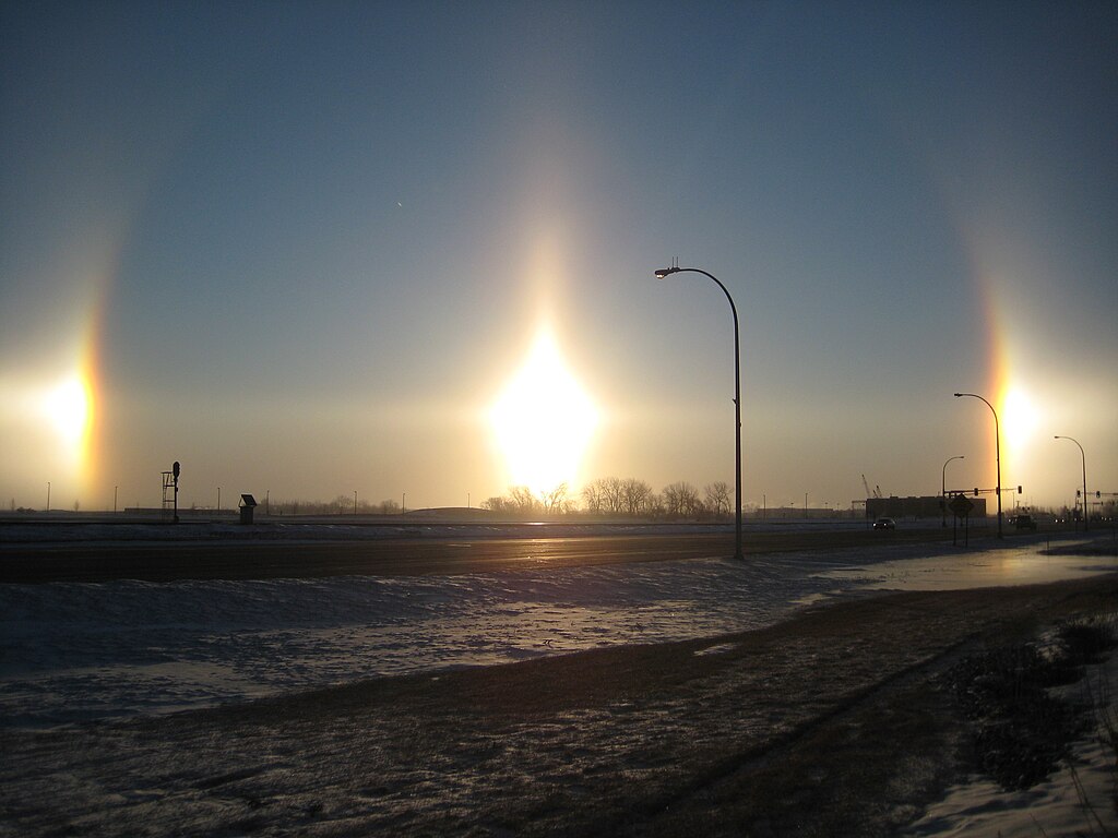 A photo of two sun dogs. The sun is in the middle of the photo. Either side of the sun are two bright spots. They sort of extend up into a faint arc.